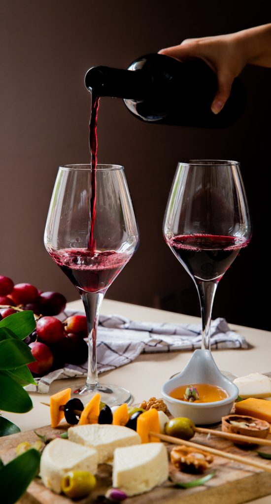 side view of woman hand pouring red wine into glass and different kinds of cheese olive walnut grape on white surface and black background